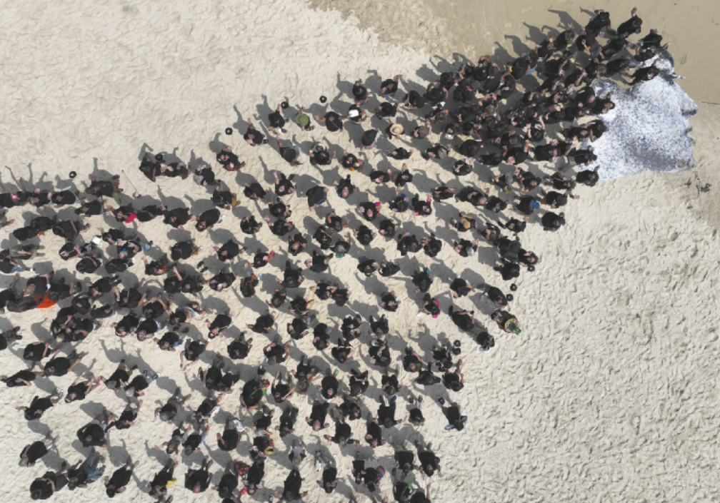 Fotografia aérea de um grande grupo de pessoas vestidas com roupas pretas em uma praia, organizadas no espaço de modo a configurar a representação dos cabelos longos de uma figura feminina impressa em grande formato, colocada sobre a areia.