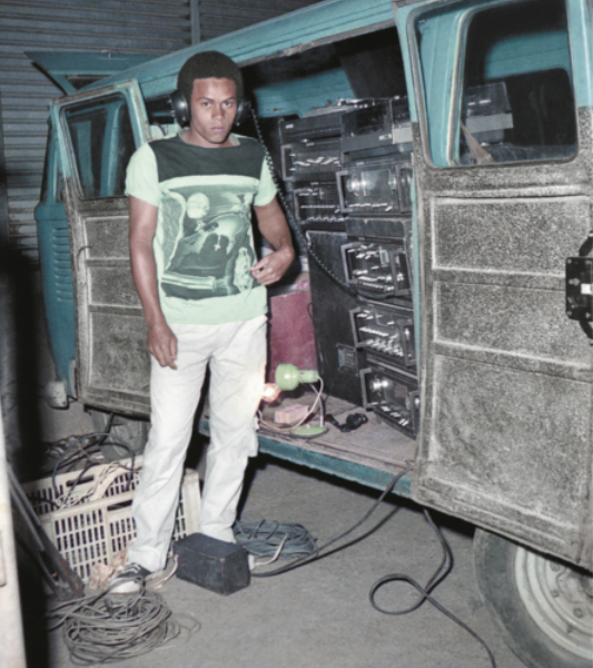 Fotografia de um homem em pé usando um fone de ouvido ao lado de uma van com a porta aberta, contendo diversas caixas de som e aparelhos de som. No chão ao lado dele há caixas de feira com fios elétricos e cabos enrolados.
