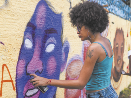 Fotografia de Nathália Ferreira. Ela é uma mulher com cabelos cacheados, usando óculos e brincos, pintando um grafite em um muro com um spray de tinta. Ela está pintando a figura de um rosto colorido.