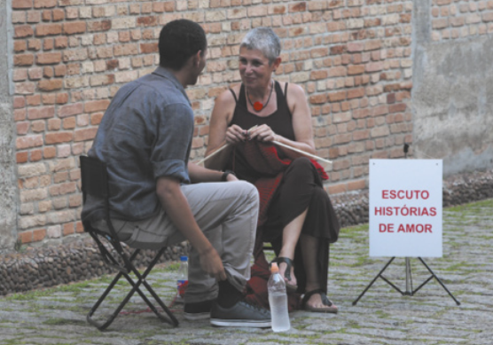 Fotografia de Ana Teixeira. Ela é uma mulher de cabelos curtos e grisalhos, sentada em uma cadeira em um espaço público, tricotando. Ela está conversando com um homem sentado em uma cadeira ao lado dela; e do outro lado dela está uma placa com a mensagem: 'Escuto histórias de amor'.