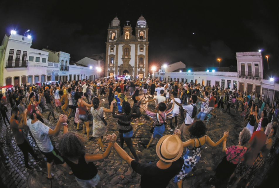 Fotografia de um grande grupo de pessoas de mãos dadas, dançando em roda em uma praça com casas, edifícios baixos e uma igreja ao fundo.