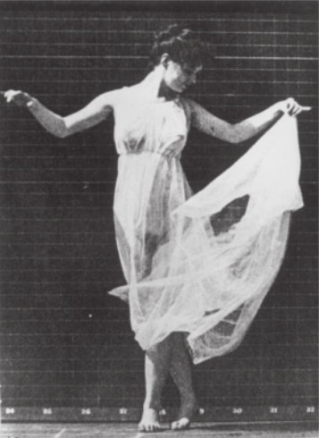 Fotografia em preto e branco de uma mulher com um vestido de tecido semitransparente, dançando em pé com uma perna cruzada sobre a outra, e com os braços abertos à altura dos ombros, segurando a saia com uma das mãos.