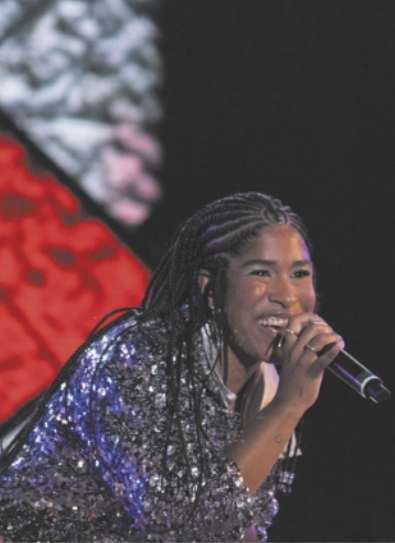 Fotografia de Drik Barbosa. Ela é uma cantora com tranças nos cabelos, vestida com um casaco brilhante, cantando com um microfone na mão.