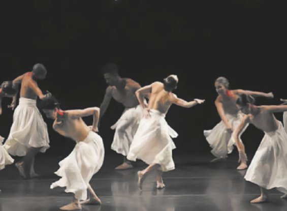 Fotografia de um grupo de homens e mulheres dançando em um palco, usando saias brancas, dançando com as pernas flexionadas e o tronco inclinado para a frente, e movimentando os braços ao lado do corpo e apoiando uma das mãos na região da lombar. Alguns deles estão em posições semelhantes à ginga da capoeira.