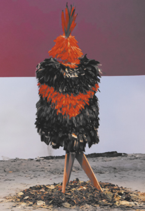 Fotografia de um manto de plumas exposto sobre um suporte de madeira, que simula o formato de um corpo em pé. O artefato está posicionado em um salão, com folhas secas no chão.