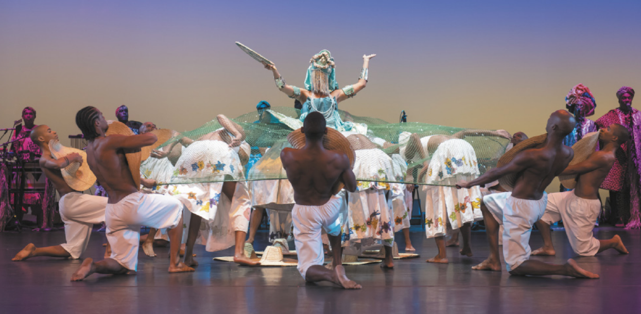 Fotografia de um grupo de dançarinos em um palco. Há homens sem camiseta ajoelhados em círculo, segurando chapéus de palha sobre o peito e as extremidades de uma rede de pesca. Dentro da rede, há bailarinas em pé, com o tronco inclinado para a frente. Ao centro, está uma bailarina com a postura ereta e os braços erguidos para o alto, vestida como Iemanjá. Ela está usando colares, braceletes e uma espécie de coroa com uma franja de tecidos que cobrem o rosto dela. Ao fundo há músicos tocando instrumentos diversos.