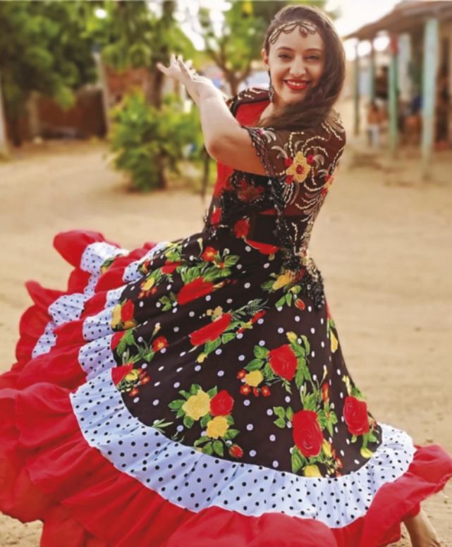 Fotografia de Marcilania Alcantara. Ela é uma mulher com cabelos longos, usando batom, brincos e uma tiara, vestida com uma saia longa e de estampa florida. Ela está dançando com os braços erguidos à altura dos ombros e as pernas afastadas, sobre um piso de areia, com arbustos e casas ao fundo.