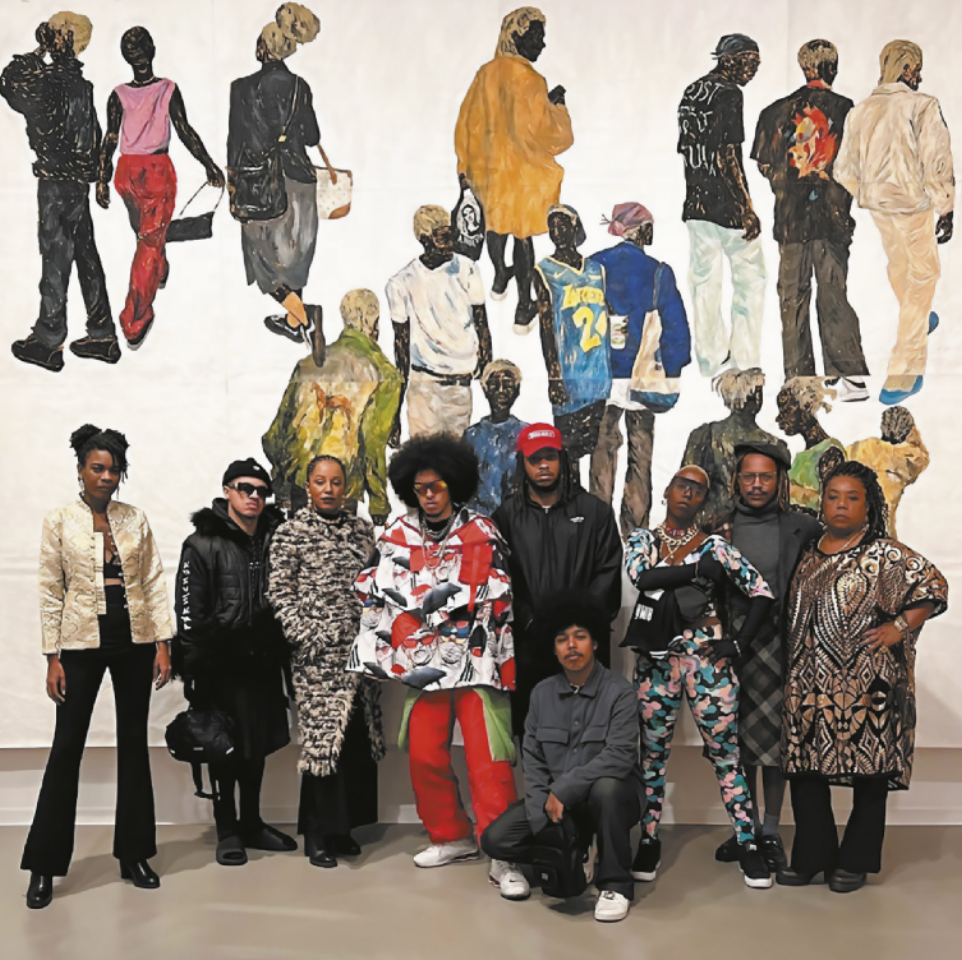 Fotografia de um grupo de pessoas em torno do artista Maxwell Alexandre. Ele é um homem com barba e tranças nos cabelos, usando um boné. As pessoas posando para a fotografia em volta dele estão vestidas com trajes de estilos diversos, com estampas e acessórios de diversas vertentes da moda contemporânea. Eles posam à frente de uma pintura de Maxwell Alexandre representando diversas pessoas negras caminhando sobre um fundo branco, vestidas com trajes de diversos tipos e estilos, além de cortes de cabelo e penteados variados, quase todas elas com os cabelos descoloridos.