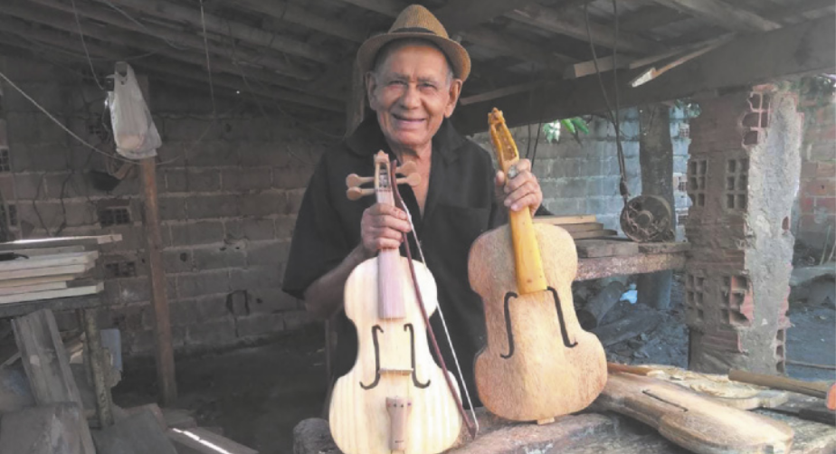 Fotografia de Nelson da Rabeca. Ele é um homem idoso de camisa e chapéu, segurando duas rabecas em processo de produção, apoiadas sobre uma mesa em sua oficina.