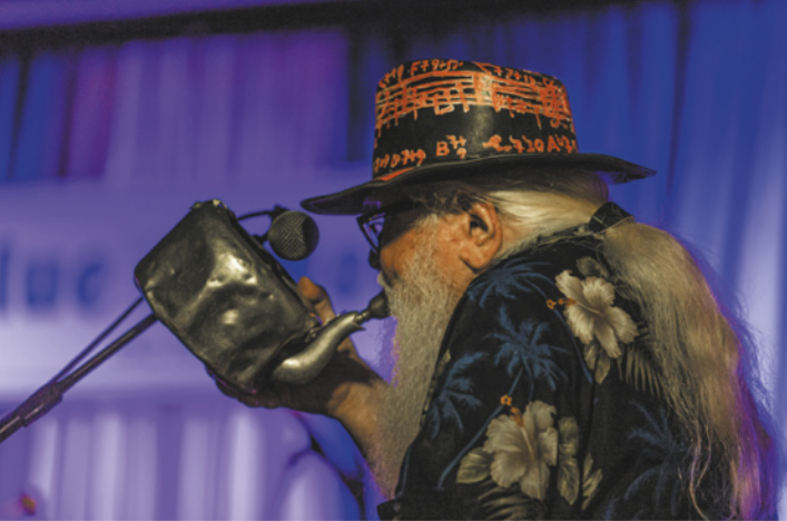 Fotografia de Hermeto Pascoal. Ele é um homem idoso, com a barba e os cabelos grisalhos e longos, de óculos, usando um chapéu e uma camisa de estampa florida. Ele está segurando uma chaleira de metal com um bocal de trompete acoplado ao bico da chaleira, tocando-a como um instrumento de sopro, diante de um microfone.