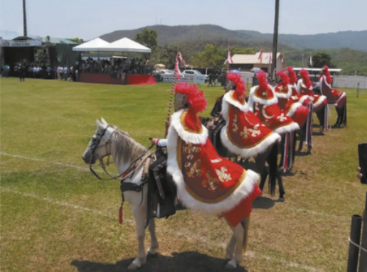 Fotografia de cavalos enfileirados em um gramado. Sobre eles estão montadas pessoas vestidas com fantasias de cavaleiros, usando mantos e capacetes com plumas no topo, segurando lanças. Ao fundo há pessoas observando, abrigadas sob tendas.
