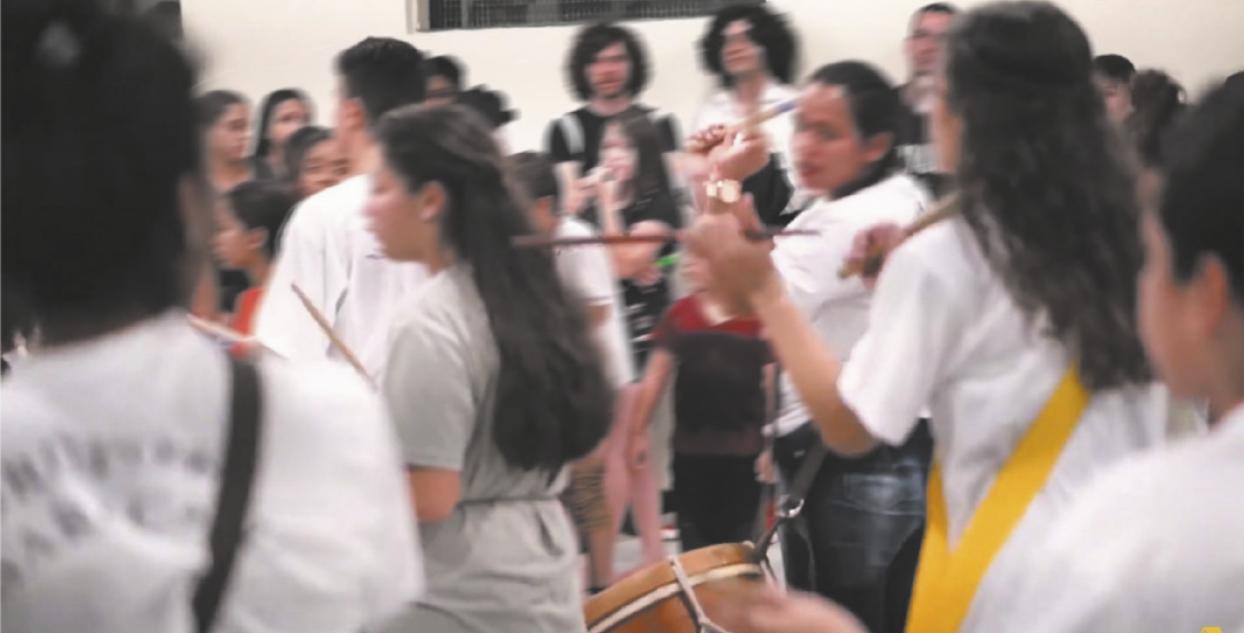 Fotografia de um grupo de jovens enfileirados tocando alfaias penduradas a tiracolo na altura das coxas, com baquetas na mão. Ao fundo há pessoas em pé assistindo.