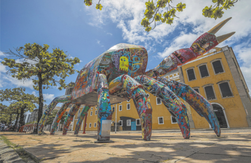 Fotografia de uma grande escultura representando um caranguejo em uma praça, em frente a um edifício baixo de arquitetura do período colonial brasileiro. A superfície da escultura é coberta por desenhos e pinturas coloridas representando figuras diversas, formas abstratas, símbolos e grafismos inspirados na linguagem visual do grafite.