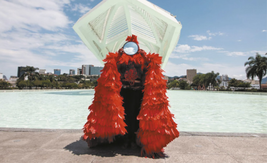 Fotografia de uma performance em que uma pessoa vestida com um manto de plumas e penas de aves segura um espelho circular em frente ao rosto. O espelho reflete a imagem do céu azul com nuvens, e a performance ocorre em uma praça com um espelho d'água em torno de um museu de arte.