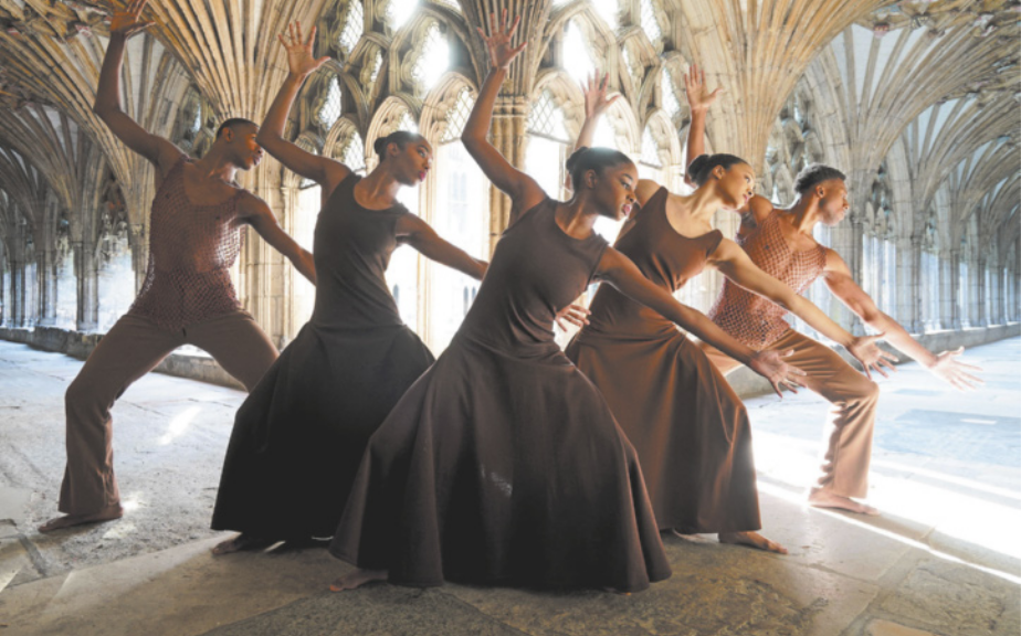Fotografia de um grupo de três bailarinas e dois bailarinos executando um movimento coreografado. Todos estão com as pernas afastadas e os joelhos flexionados, inclinando o corpo para o lado esquerdo, com o braço esquerdo estendido em diagonal para baixo, e o braço direito erguido para o alto. Eles dançam em um corredor com arcadas rebuscadas e ornamentadas de uma catedral de arquitetura gótica.