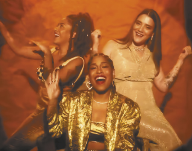 Fotografia de três mulheres dançando e cantando: elas são Drik Barbosa, ao centro, usando uma jaqueta dourada, com os cabelos presos, batom e brincos com a forma de corações; e ao lado dela, Cynthia Luz e Lourena estão dançando sorridentes, usando vestidos, gesticulando com as mãos.
