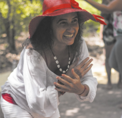 Fotografia de Lilian Pacheco. Ela é uma mulher de cabelos longos, usando um colar de conchas e um chapéu enfeitado com um laço. Ela está sorrindo com as mãos cruzadas à frente do peito, com o tronco inclinado para a frente.
