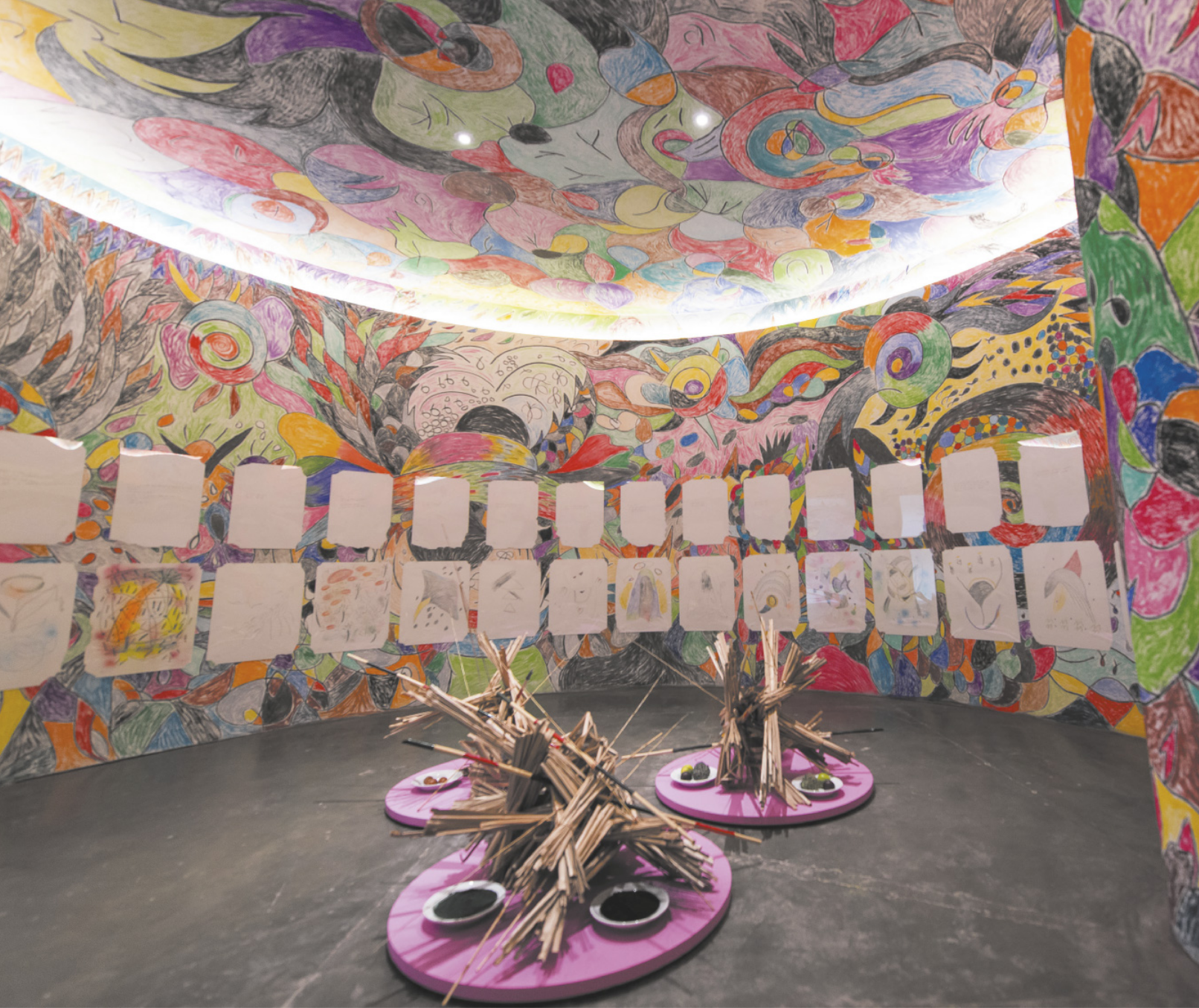 Fotografia de instalação artística em um ambiente expositivo circular, com desenhos coloridos abstratos de formas curvas e arredondadas ocupando o teto e as paredes, além de desenhos em folhas brancas expostos nas paredes e três esculturas expostas sobre pedestais circulares no chão, feitas com varetas de madeira aglomeradas e justapostas, ao lado de pratos com alimentos.