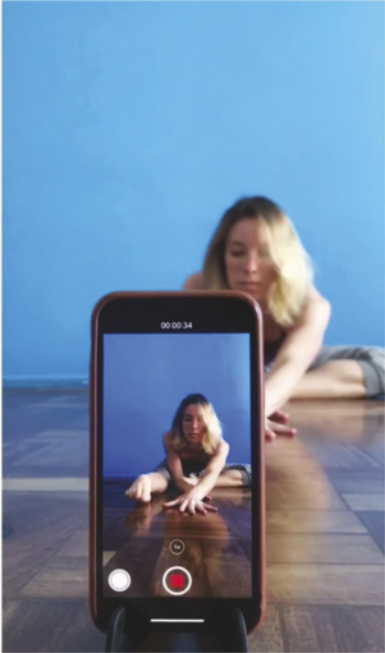 Fotografia de um smartphone apoiado em um suporte no chão, filmando uma mulher sentada no chão com uma das pernas estendida à frente e a outra flexionada. Ela inclina o tronco para a frente, estendendo os braços no chão na mesma direção da perna.