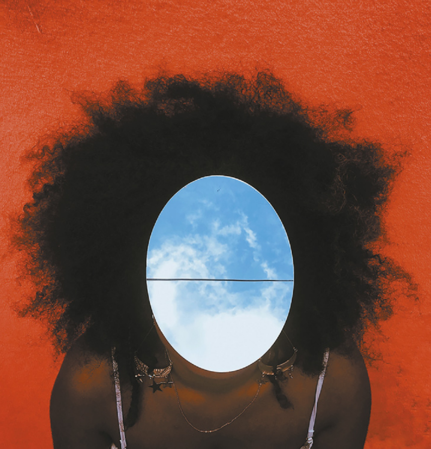 Fotografia com manipulação digital do retrato de uma mulher de cabelos cacheados e volumosos. Sobre o rosto dela, foi inserida uma elipse que contém a imagem de um céu azul com nuvens.