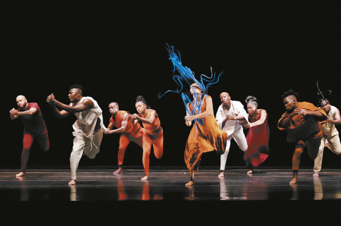 Fotografia de um grupo de dançarinos em um palco. Eles estão enfileirados realizando um mesmo movimento, pisando com um só pé no chão, com o corpo inclinado para a frente e as mãos unidas à frente do peito. Ao centro, uma dançarina dança agitando os cabelos com tranças longas e esvoaçantes pintadas de azul.