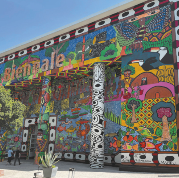 Fotografia de pintura de mural sobre a fachada de um edifício com colunas na entrada. A pintura representa diversas figuras de animais, elementos da natureza, artefatos e símbolos das culturas indígenas, com cores vivas e muitos grafismos geométricos e abstratos em meio às figuras.