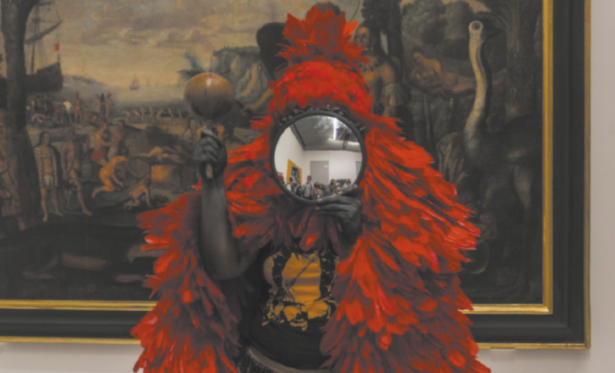 Fotografia de uma mulher indígena vestida com um manto de plumas, com um espelho circular cobrindo o rosto, segurando um chocalho. Ela está em um espaço expositivo, à frente de uma pintura representando indígenas em uma paisagem, com navios colonizadores no mar ao fundo.