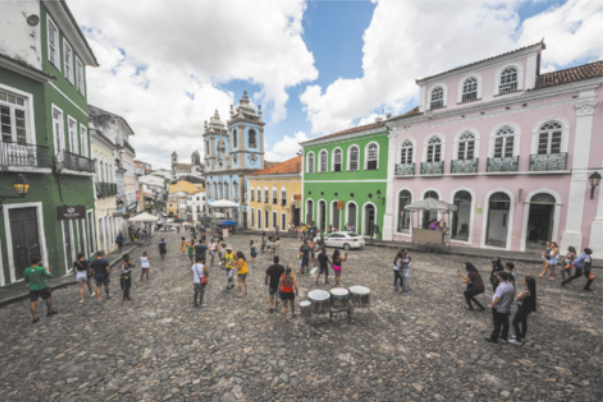 Fotografia de uma praça em uma via pública com piso de pedra, com edifícios baixos de arquitetura do período colonial brasileiro e uma igreja. Há um conjunto de tambores no centro da praça e pessoas caminhando.