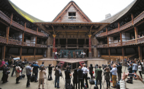 Fotografia do interior de um teatro com estruturas de madeira e arquibancadas laterais distribuídas em três níveis. Ao centro, há um palco coberto por um telhado, com um andar superior em formato triangular. Um grupo de atores está no palco encenando, enquanto diversas pessoas assistem em pé na plateia; entre elas, muitos fotógrafos registram a apresentação.