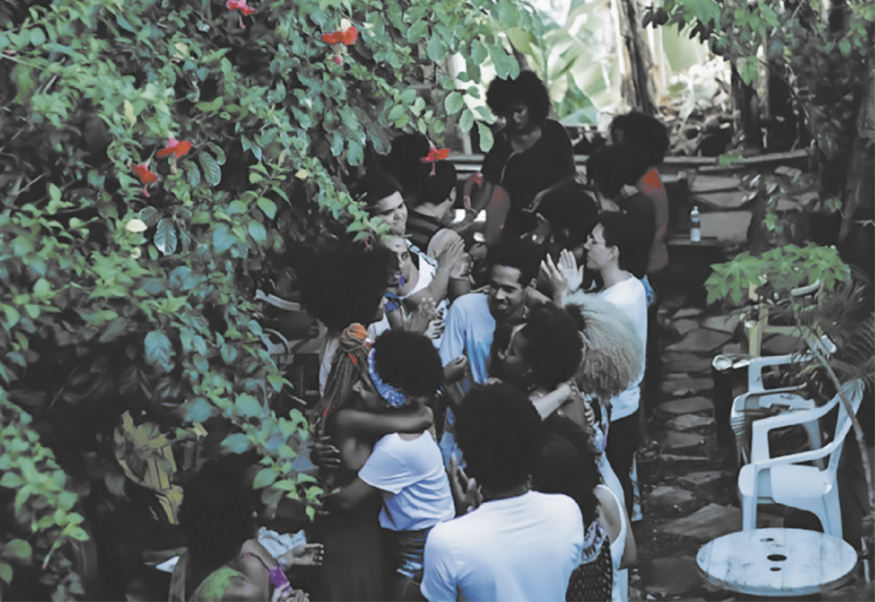 Fotografia de um grupo de jovens conversando, se abraçando e batendo palmas em um jardim com cadeiras e mesas.