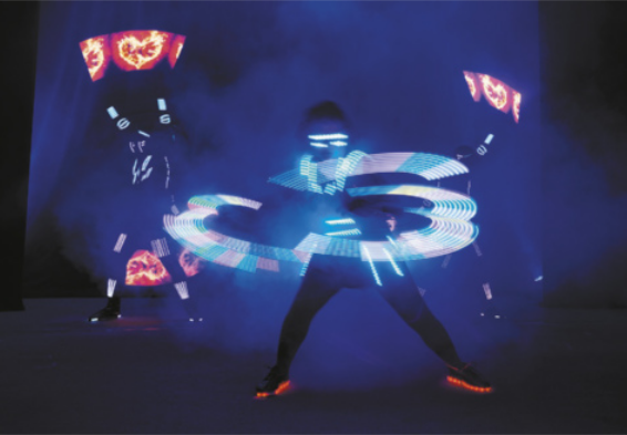 Fotografia de três pessoas em um palco escuro, com fumaça cênica e luz colorida, usando roupas escuras com detalhes luminosos. Dois deles estão erguendo objetos luminosos para o alto, enquanto um deles, à frente, movimenta os braços de modo que se formam desenhos luminosos no ar.