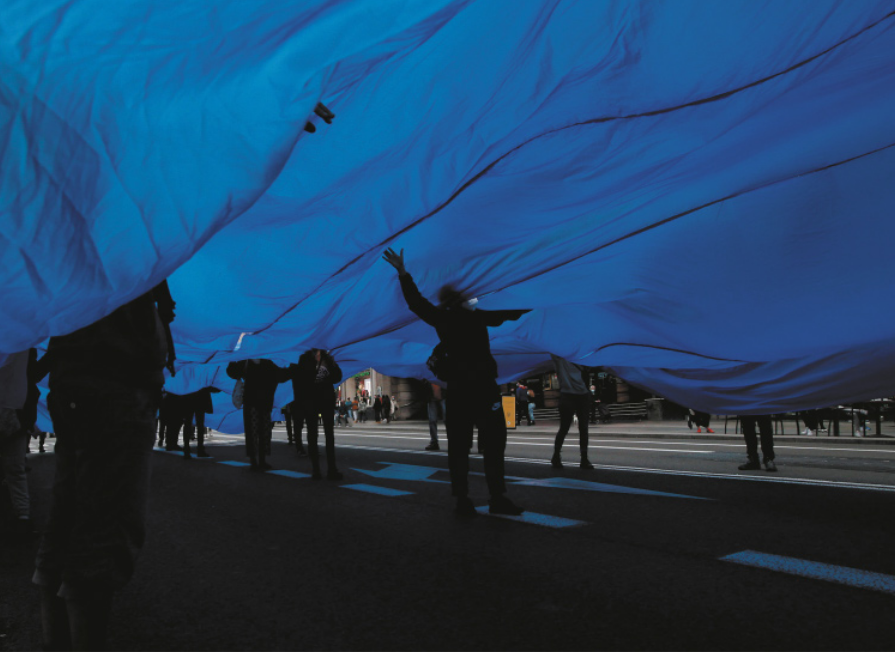 Fotografia de pessoas em protesto em uma via pública, realizando performance em que carregam uma imensa faixa de tecido azul sobre o corpo, à altura dos ombros, com as cabeças posicionadas acima do tecido, passando por orifícios espalhados pela extensão dele.