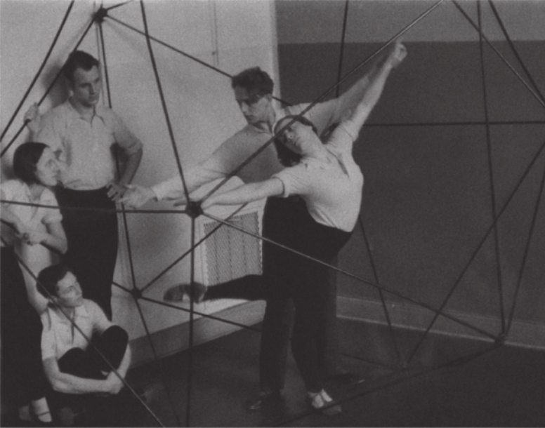 Fotografia de um grupo de estudantes de dança. Um homem e uma mulher estão realizando movimentos com os braços abertos, no espaço interno de uma estrutura de hastes metálicas que configura uma esfera vazada com faces triangulares. O homem está atrás da mulher, segurando uma das mãos dela no alto, e ela executa uma pose de dança, com um dos pés erguidos para trás, inclinando a cabeça para trás e para o lado, acompanhando a direção do outro braço. Dois homens e uma mulher estão ao lado deles, observando.