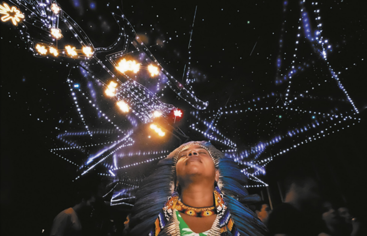 Fotografia de uma mulher indígena usando um cocar de penas, um colar e outros adereços, com os olhos fechados e a cabeça voltada para cima. Ao fundo há uma instalação de linhas pontilhadas luminosas pendendo de um teto com um céu estrelado.