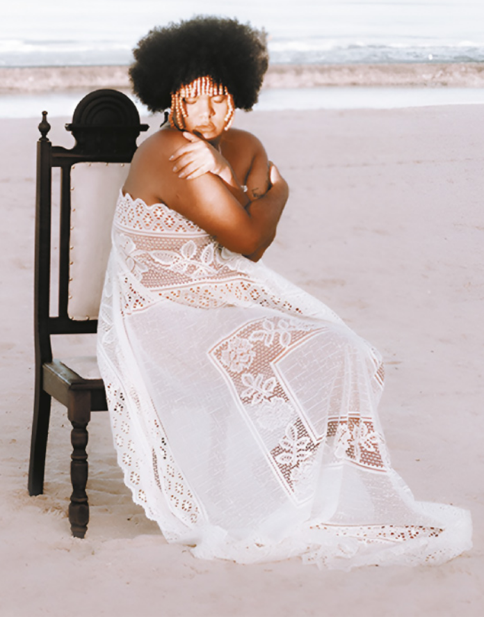 Fotografia de Ariel Castiel. Ela é uma mulher com cabelos cacheados e adereços de contas amarrados aos cabelos na região da franja. Ela está usando um vestido de rendas, sentada em uma cadeira sobre a areia de uma praia, com os braços cruzados, abraçando os próprios ombros.