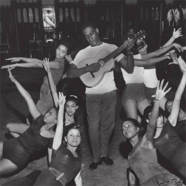 Fotografia em preto e branco de um homem em pé tocando violão rodeado por um grupo de dançarinas executando poses em pé e com o corpo deitado próximo ao chão, com os braços para o alto.
