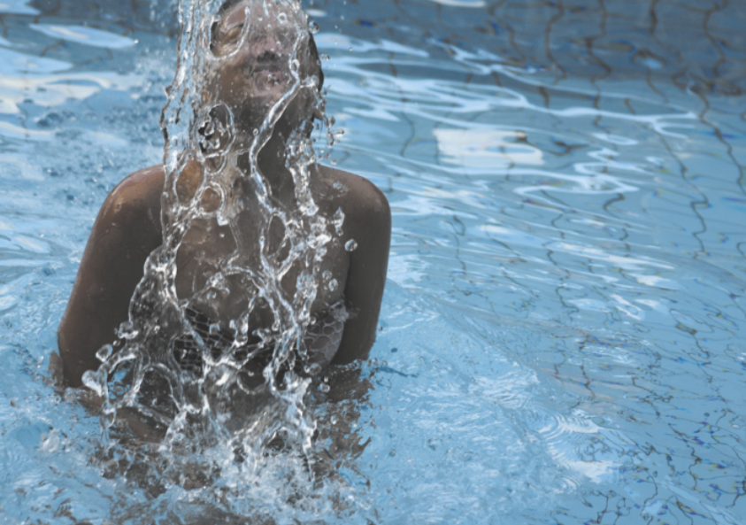 Fotografia de um atleta em uma piscina, usando maiô, saltando com a cabeça para fora da água. Grandes gotas voam à frente do seu rosto, enquanto a superfície agitada da piscina reflete luzes e distorce as linhas dos azulejos no fundo.