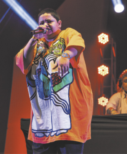 Fotografia de Jéssica Caitano. Ela é uma mulher com cabelos curtos, usando uma camiseta longa, cantando em um palco com um microfone na mão.