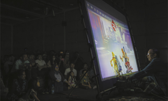 Fotografia de uma plateia assistindo a um espetáculo de fantoches em uma sala escura, diante de uma tela iluminada semitransparente, atrás da qual está sentado um homem, que manipula dois fantoches próximos à tela.