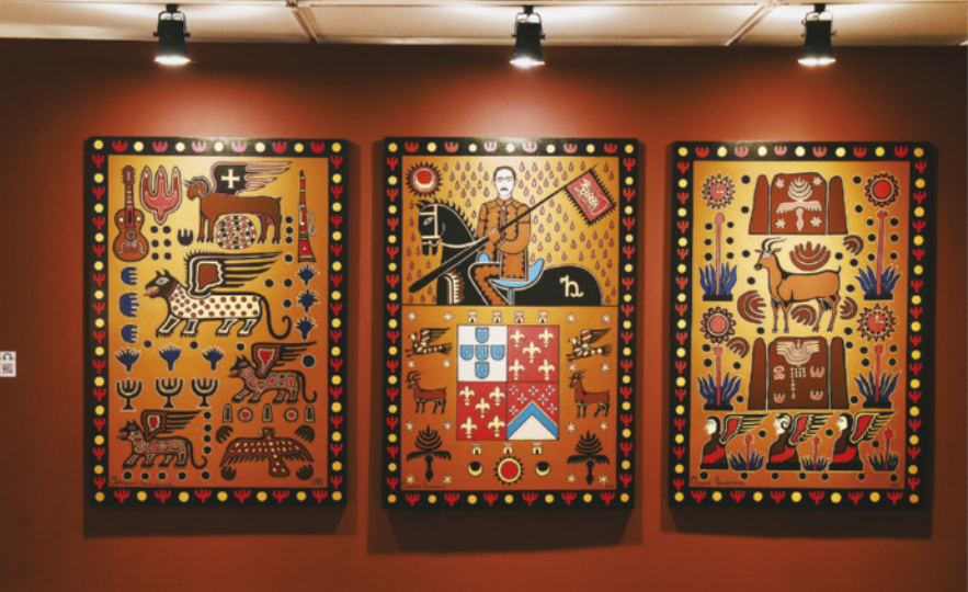 Fotografia de três pinturas em uma parede de espaço expositivo. Elas representam diversas figuras simbólicas, como Lampião andando a cavalo, animais, seres míticos, elementos da natureza, instrumentos musicais, entre outros elementos representativos da cultura nordestina, pintados segundo uma linguagem visual estilizada e sintética, com contrastes fortes, cores vivas e grafismos decorativos tradicionais.