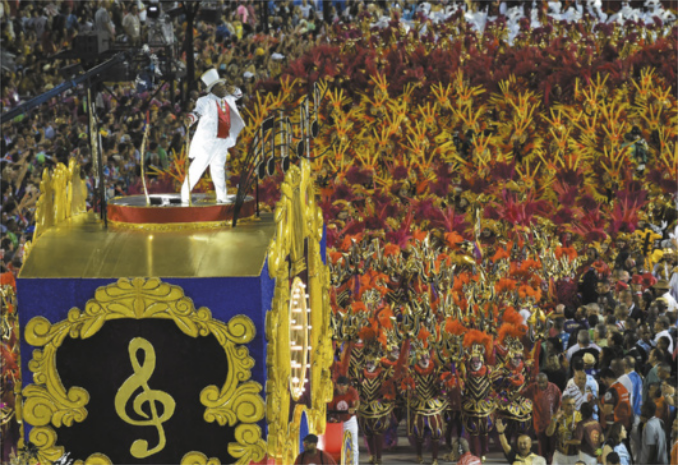 Fotografia de uma multidão de foliões em um desfile de escolas de samba no carnaval. Eles caminham, uniformizados com trajes brilhantes e enfeitados com plumas, atrás de um trio elétrico com ornamentos dourados e o símbolo de uma clave de sol, no topo do qual está em pé um homem de terno branco e cartola.