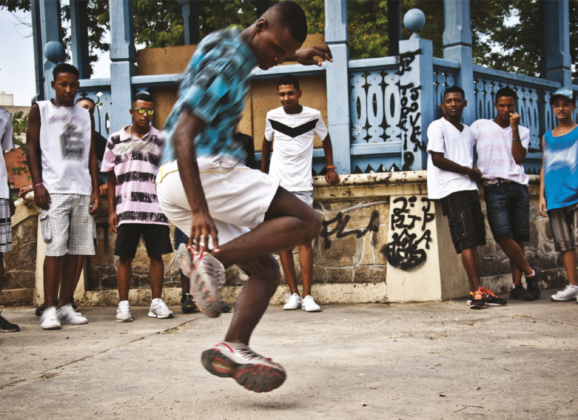 Fotografia de um homem ao centro de uma roda de jovens em uma praça, executando um passo de dança com o corpo no ar, flexionando os joelhos em um salto e tocando um dos tênis com a mão. Ao fundo, jovens em pé observam a apresentação do homem ao centro, apoiados em um coreto.