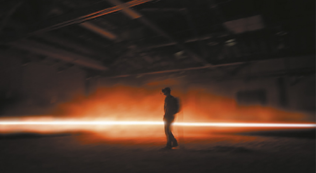 Fotografia de um homem usando um óculos de realidade virtual em um espaço expositivo escuro com um feixe de luz horizontal passando pelo espaço, com uma fumaça cênica que refrata a luz com cores avermelhadas.