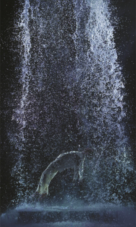 Fotografia de um homem saltando para trás, com o corpo inclinado no ar sob a queda d’água de uma cachoeira iluminada, tendo ao fundo um ambiente escuro.