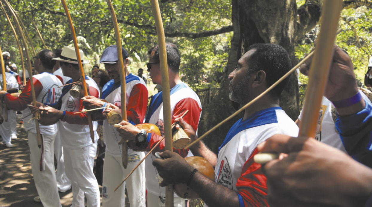 Fotografia de um grupo de homens tocando berimbaus, enfileirados e uniformizados, em um parque arborizado à beira de um rio.