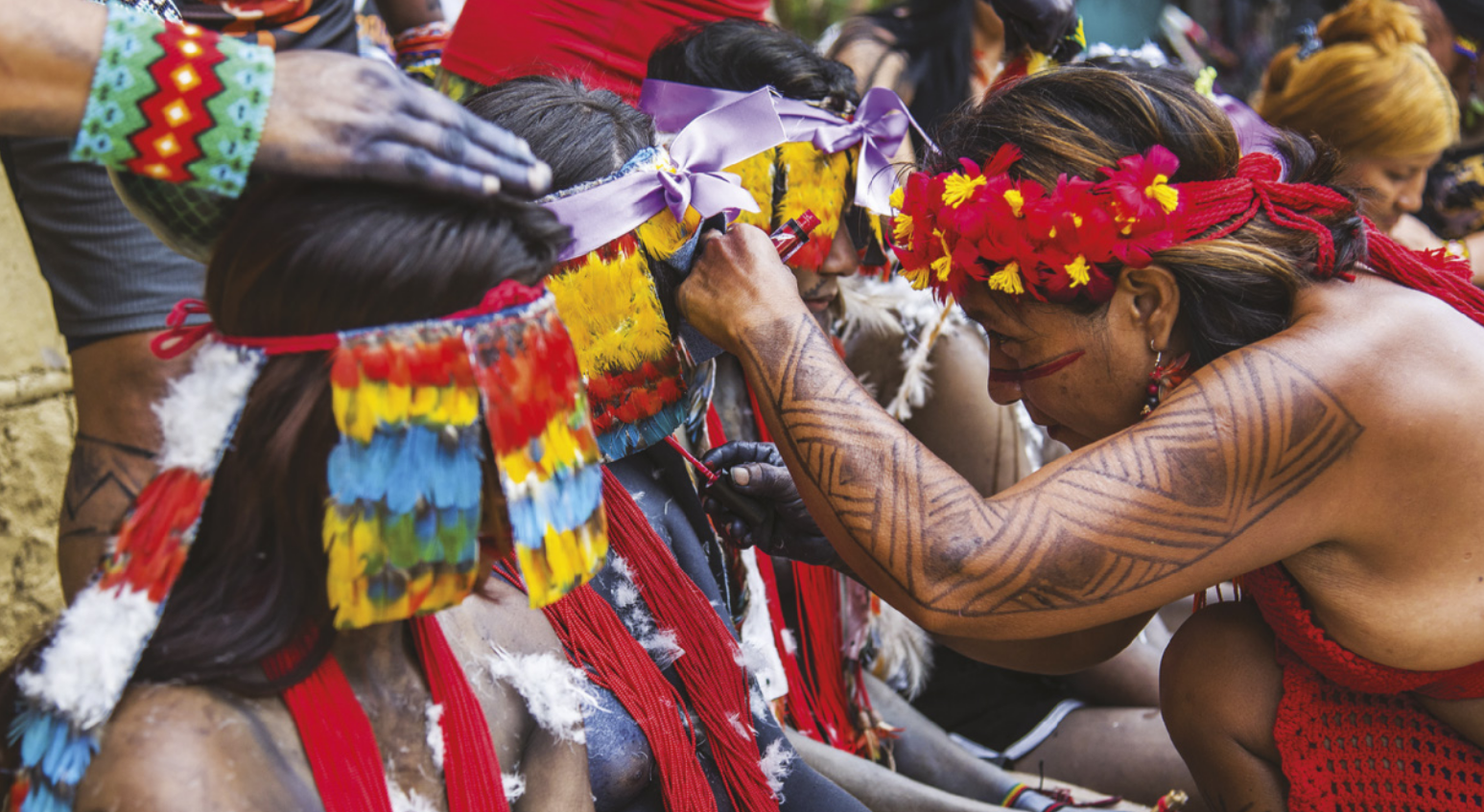 Fotografia de mulheres indígenas sentadas enfileiradas, usando cocares de penas coloridas que lhes cobrem os rostos, colares e pintura corporal. Uma mulher com pinturas corporais está pintando o rosto de uma delas.
