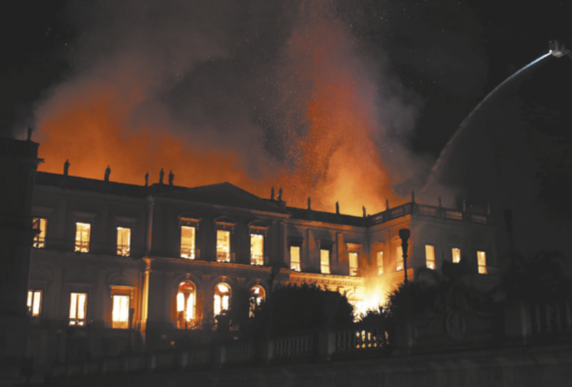 Fotografia da fachada de um edifício de arquitetura neoclássica à noite, com o interior em chamas e fumaça saindo pelo telhado.
