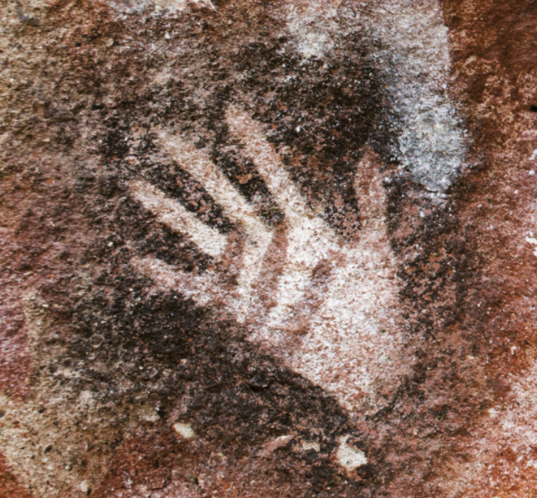 Detalhe de pintura rupestre com figura de mão humana de cor branca sobre um fundo escuro.