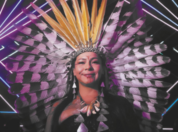 Fotografia de Daiara Tukano. Ela é uma mulher indígena usando um cocar de penas, brincos, um colar e pinturas no rosto. Ao fundo há linhas em diagonal com luzes coloridas, que fazem parte de uma instalação artística.