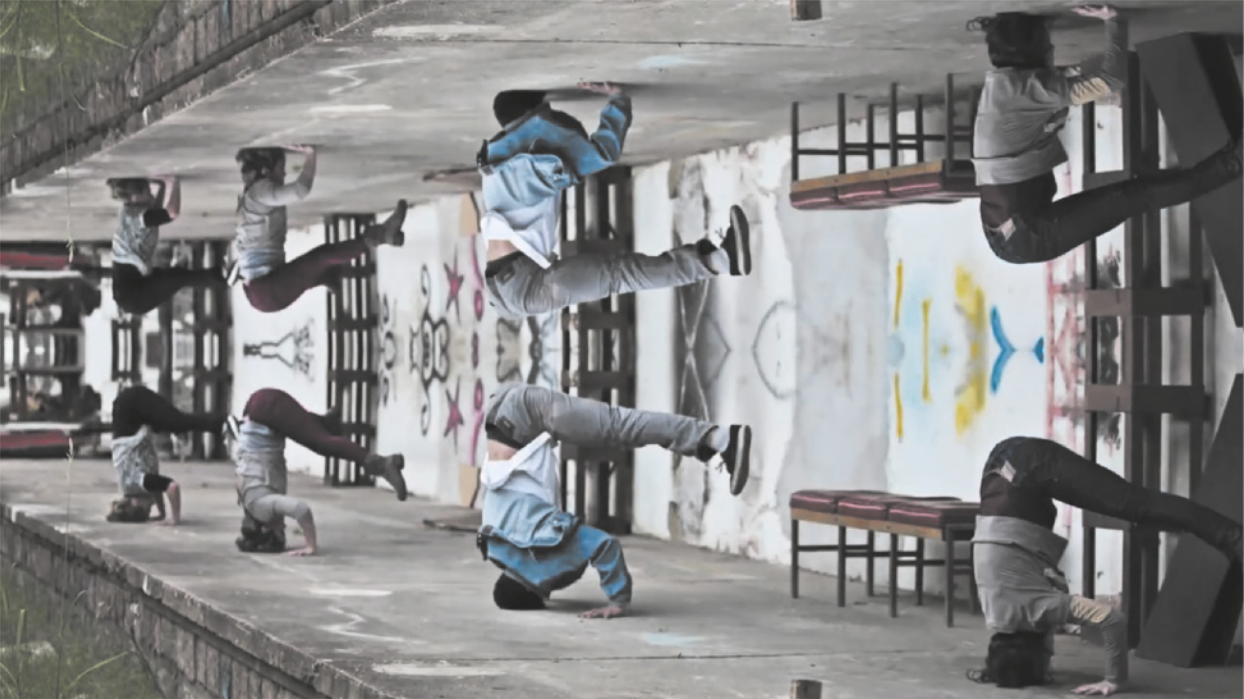 Fotografia com manipulação digital mostrando pessoas realizando paradas de cabeça em uma estação de trem. Elas estão enfileiradas, apoiadas no chão com a cabeça e as mãos, com os quadris elevados e as pernas estendidas e afastadas na horizontal. A imagem foi espelhada verticalmente, criando a ilusão de pessoas idênticas penduradas no teto da estação, reproduzindo exatamente as mesmas posições das figuras no chão.